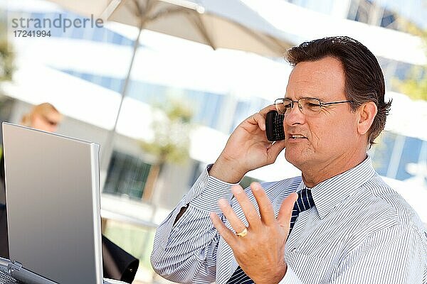 Gutaussehender Geschäftsmann  der mit seinem Handy telefoniert und im Freien am Laptop arbeitet