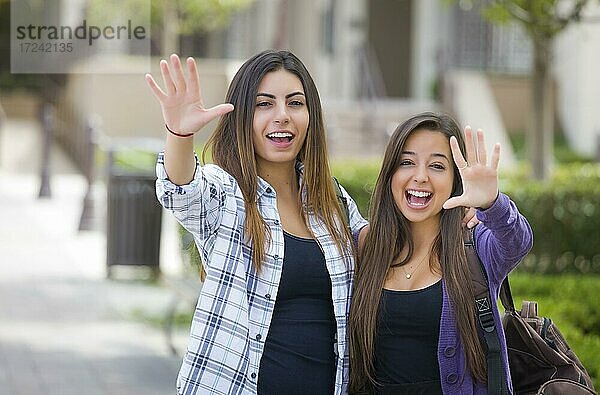 Porträt von zwei attraktiven multiethnischen Studentinnen winken und tragen Rucksäcke auf dem Schulcampus