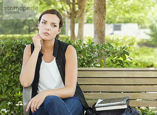 Melancholische junge erwachsene Frau sitzt auf Bank neben Bücher und Rucksack
