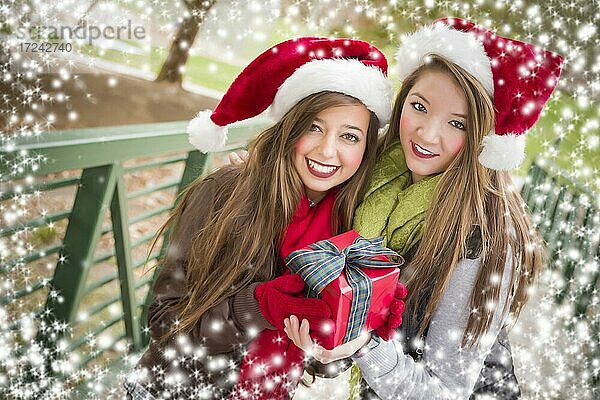 Zwei attraktive festlich gekleidete lächelnde multiethnische Frauen tragen Weihnachten Santa Hüte halten eine Schleife eingewickelt Geschenk außerhalb mit Schneeflocken