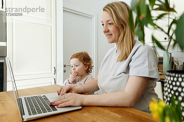 Lächelnde Unternehmerin  die mit ihrer Tochter am Tisch in der Küche an einem Laptop arbeitet