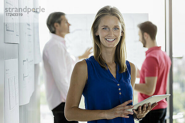 Junge Geschäftsfrau hält digitales Tablet mit männlichen Kollegen im Hintergrund im Büro