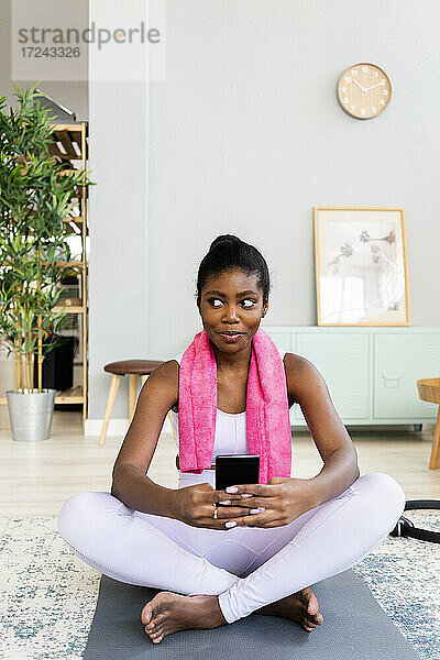 Junge Frau benutzt ein Mobiltelefon  während sie zu Hause auf einer Gymnastikmatte sitzt