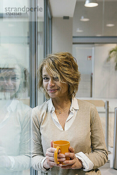 Reife Geschäftsfrau mit blondem Haar  die einen Kaffee trinkt  während sie am Fenster im Büro steht