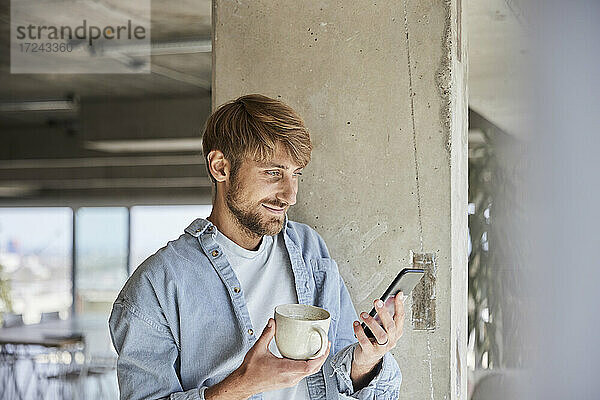 Blonder Mann mit Kaffeetasse und Handy an Säule gelehnt im Loft