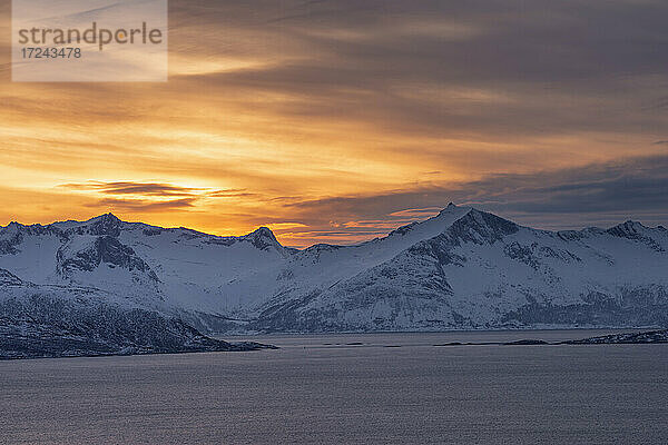 Norwegen  Tromso  Kvaloya  Dramatischer Himmel über dem schneebedeckten Berg Senja Island von der Insel Kvaloya aus gesehen bei Sonnenuntergang