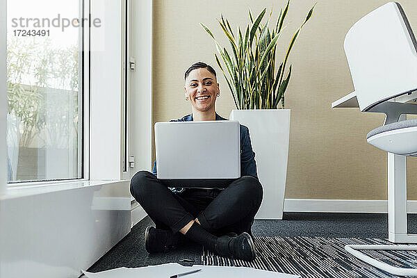 Lächelnde Geschäftsfrau mit Laptop auf dem Schoß sitzt auf dem Boden im Büro