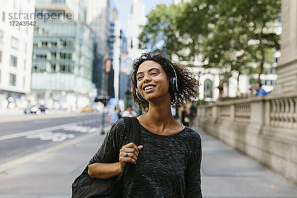 Lächelnde Frau  die wegschaut  während sie auf einem Fußweg in der Stadt über Kopfhörer Musik hört
