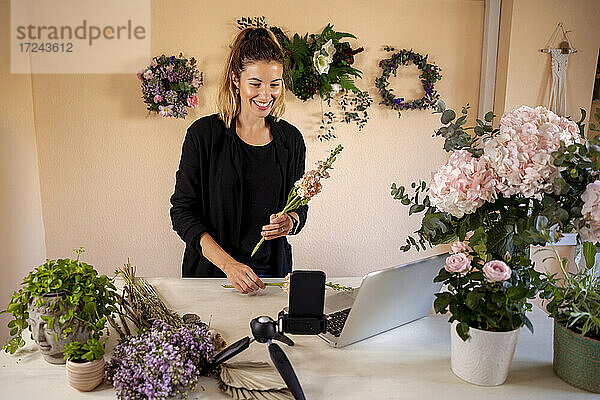Lächelnde Floristin  die eine Blume hält  während sie einen Videoanruf in einer Werkstatt tätigt