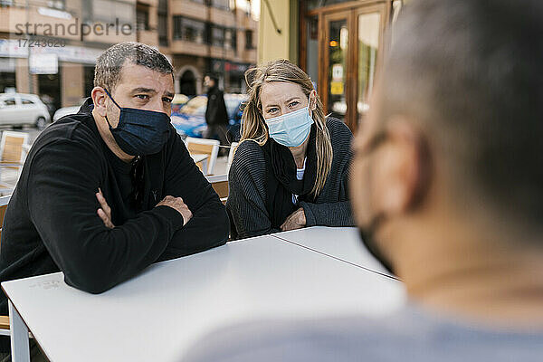 Männliche und weibliche Freunde tragen Gesichtsmasken  während sie auf der Terrasse einer Bar sitzen
