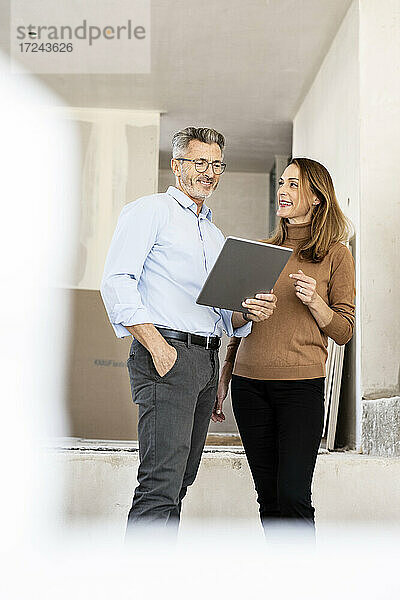 Lächelnde Kollegin  die einen männlichen Architekten betrachtet  der ein digitales Tablet auf einer Baustelle hält