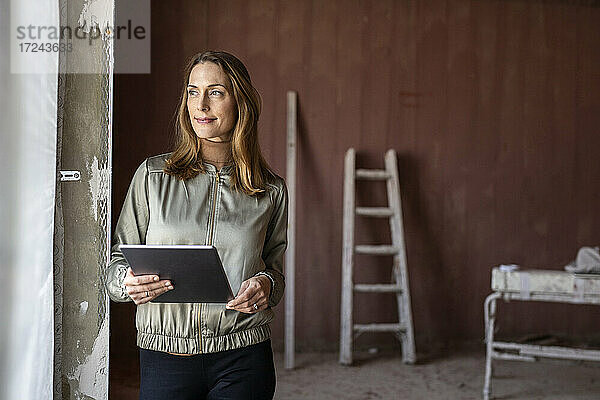 Architektin schaut weg  während sie ein digitales Tablet in der Nähe eines Fensters auf einer Baustelle hält