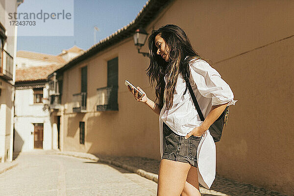 Junge Frau  die ein Mobiltelefon benutzt  während sie mit Rucksack an einem sonnigen Tag steht