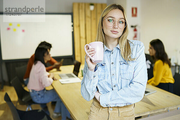 Blondhaarige kreative Geschäftsfrau  die einen Kaffeebecher hält und sich an einen Schreibtisch lehnt