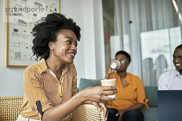 Lächelnde weibliche Fachkraft hält Einweg-Kaffeebecher im Coworking-Büro