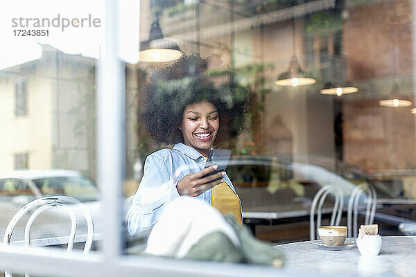 Lächelnde Geschäftsfrau  die ein Smartphone in einem Café benutzt  gesehen durch Glas