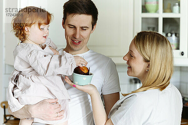 Lächelnde Frau  die ihrer Tochter von ihrem Mann in der Küche ein Törtchen schenkt