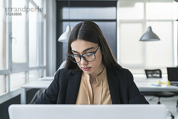 Geschäftsfrau mit Brille  die nach unten schaut  während sie im Büro vor einem Laptop sitzt