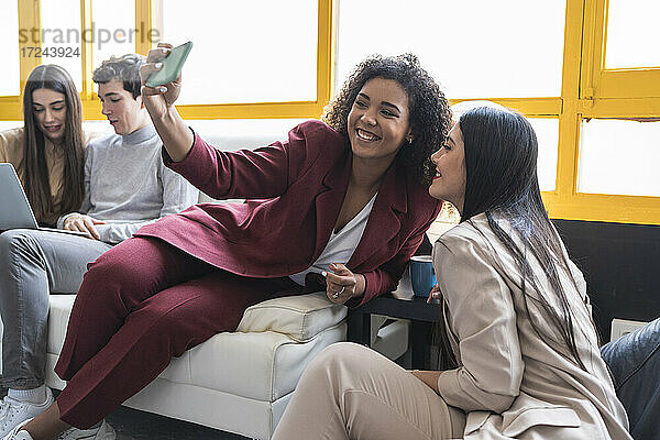 Lächelnde Unternehmerinnen  die ein Selfie mit ihrem Smartphone machen  während ihre Kollegen im Coworking-Büro arbeiten