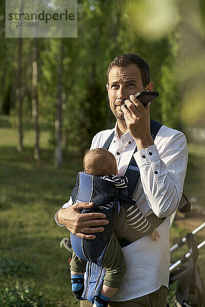 Vater trägt seinen Sohn in der Babytrage  während er im Park mit dem Handy telefoniert
