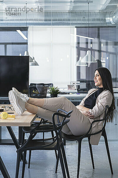 Lächelnde Geschäftsfrau mit hochgelegten Füßen  die wegschaut  während sie am Schreibtisch in einem Coworking-Büro sitzt