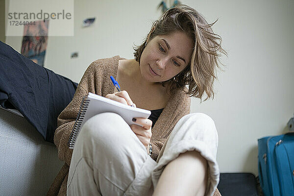 Frau mit braunen Haaren  die zu Hause vor dem Bett sitzt und auf ein Buch zeichnet