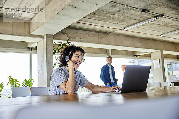 Lächelnde Frau mit Kopfhörern und Laptop mit ihrem Freund im Hintergrund in ihrer Wohnung