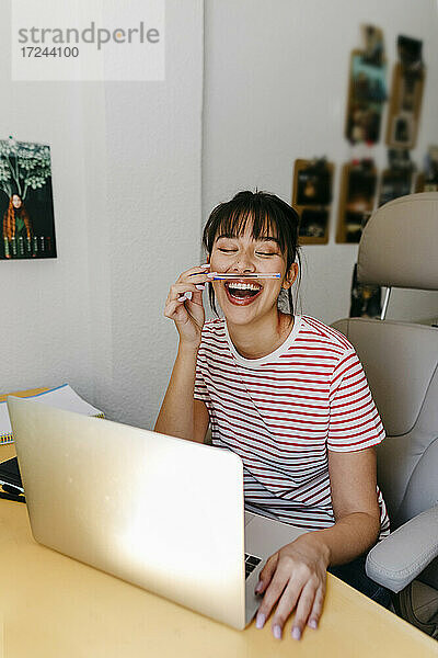 Fröhliche Frau  die mit einem Stift spielt  während sie mit einem Laptop im Büro zu Hause sitzt