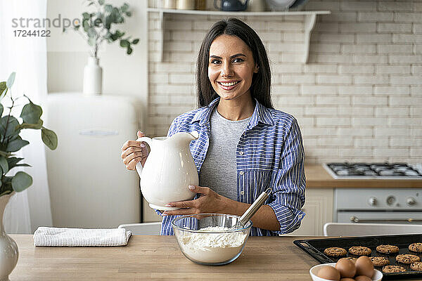 Lächelnde Frau hält Krug in der Nähe von Mehl in der Küche zu Hause