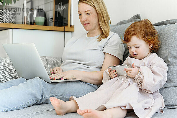 Mädchen  das ein Smartphone benutzt  sitzt neben der Mutter  die zu Hause am Laptop arbeitet