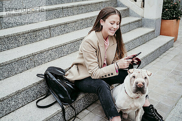 Schöne Frau mit Shar-Pei-Hund  die auf einer Treppe sitzend ein Mobiltelefon benutzt