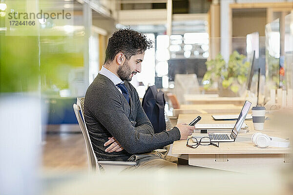 Porträt eines jungen Mannes mit Krawatte  der am Schreibtisch vor einem Laptop sitzt und auf sein Mobiltelefon schaut