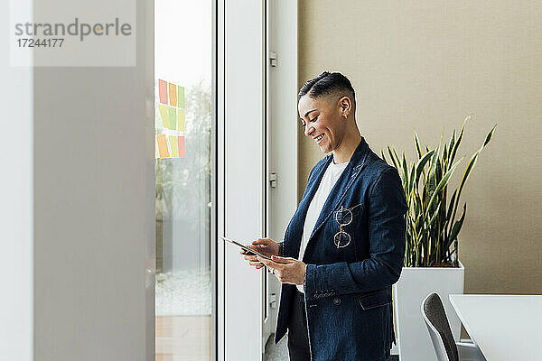 Lächelnder Unternehmer mit digitalem Tablet am Fenster im Büro