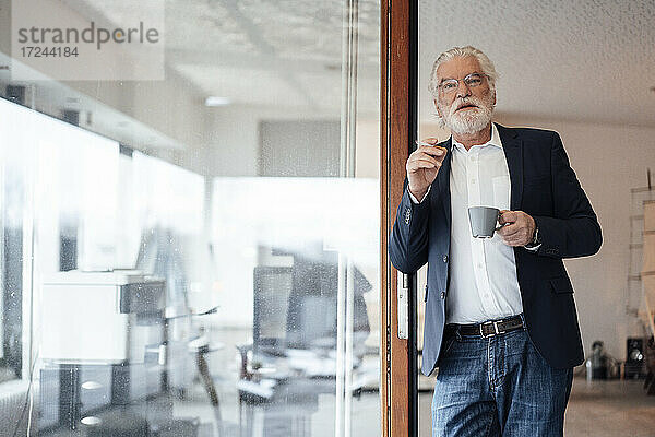 Älterer Geschäftsmann hält Zigarette mit Kaffeetasse  während er sich an eine Glaswand im Büro lehnt