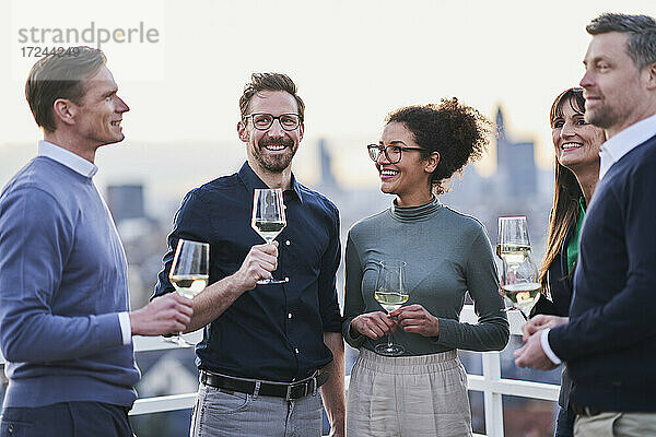Lächelnde männliche und weibliche Unternehmer stehen mit Weingläsern auf der Terrasse eines Gebäudes