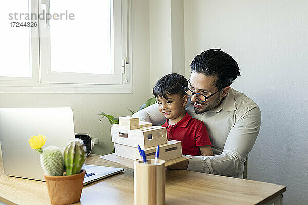 Männlicher Architekt  der ein Modell während eines Videogesprächs über einen Laptop zeigt  während er mit einem Jungen zu Hause im Büro sitzt
