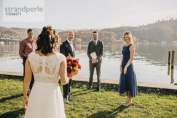 Bräutigam stehend mit Hochzeitsgästen mit Blick auf die Braut vor dem See auf Hochzeitsfeier