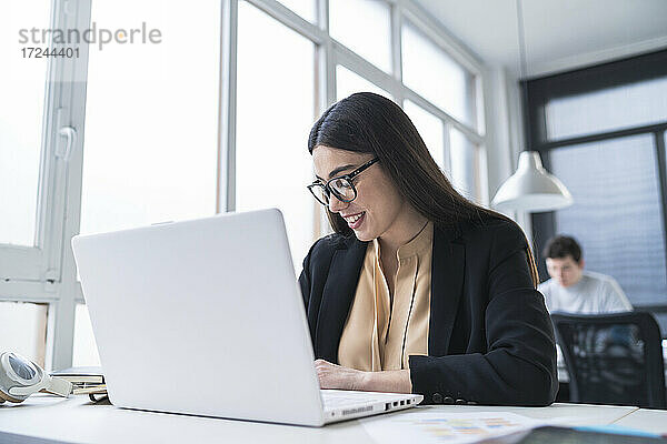 Eine Unternehmerin lächelt  während sie vor einem Laptop sitzt und ein männlicher Kollege im Hintergrund in einem Coworking-Büro arbeitet