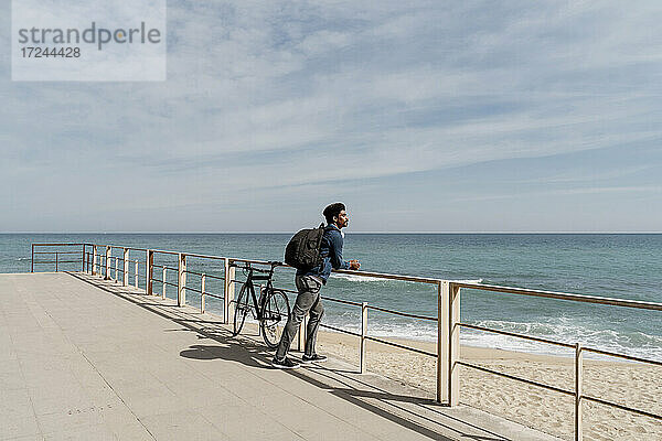 Mittlerer Erwachsener  der einen Rucksack trägt  während er sich mit dem Fahrrad am Strand auf das Geländer stützt  an einem sonnigen Tag