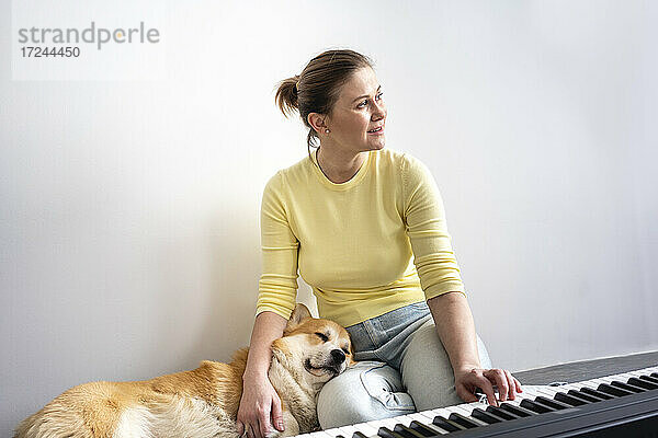 Lächelnde Frau  die wegschaut  während ihr Hund auf dem Schoß vor dem Klavier zu Hause schläft