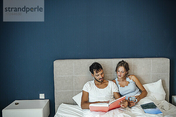 Junges Paar liest ein Buch  während es zu Hause auf dem Bett sitzt