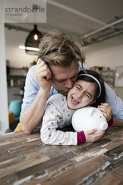 Vater küsst fröhliche Tochter mit Sparschwein auf dem Tisch zu Hause