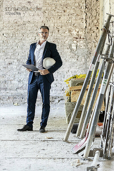 Männlicher Architekt mit Schutzhelm  der ein digitales Tablet hält  während er auf einer Baustelle steht