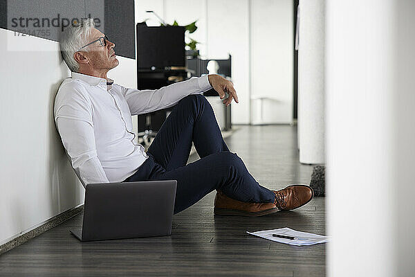 Müder Geschäftsmann sitzt mit Laptop auf dem Boden im Büro