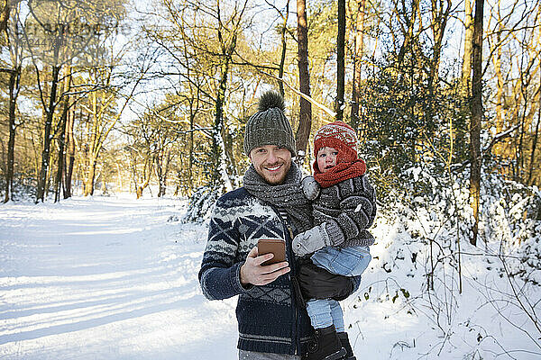 Vater mit Smartphone trägt Sohn im Winter durch den Schnee
