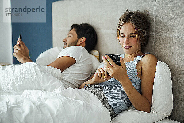 Junges Paar benutzt ein Smartphone  während es zu Hause auf dem Bett liegt