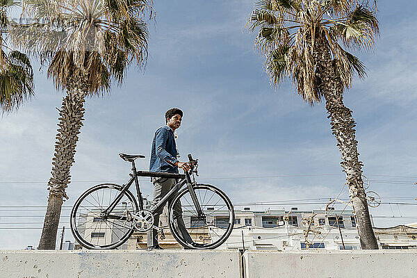 Mann mit Fahrrad  der an einem sonnigen Tag auf einer Stützmauer spazieren geht