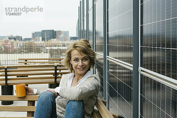 Lächelnde Geschäftsfrau mit Kaffeetasse an der Wand sitzend auf der Terrasse