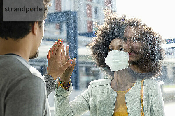 Frau mit Gesichtsschutzmaske sieht männlichen Freund durch Glaswand an