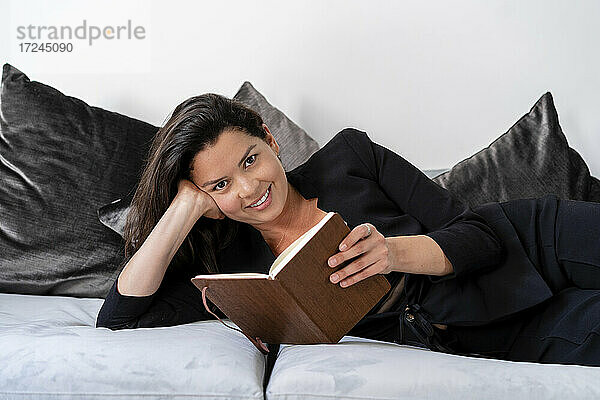 Lächelnde Frau mit Buch auf Sofa im Wohnzimmer liegend